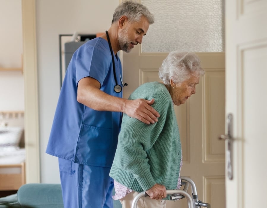 Elderly patient Mary using a walker as a male caregiver guides her.